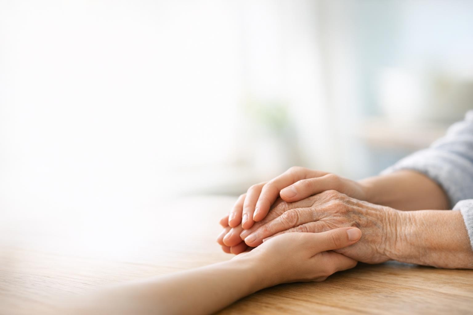 A younger person's hands gently holding an older adult's hands in soft light